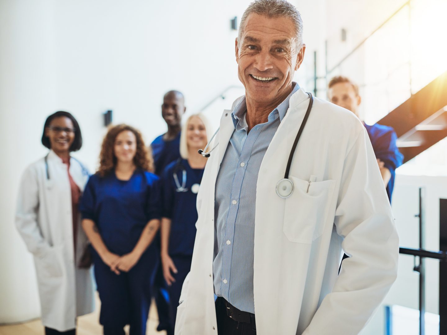 Smiling mature male doctor standing in a hospital corridor with a diverse group of staff standing behind him