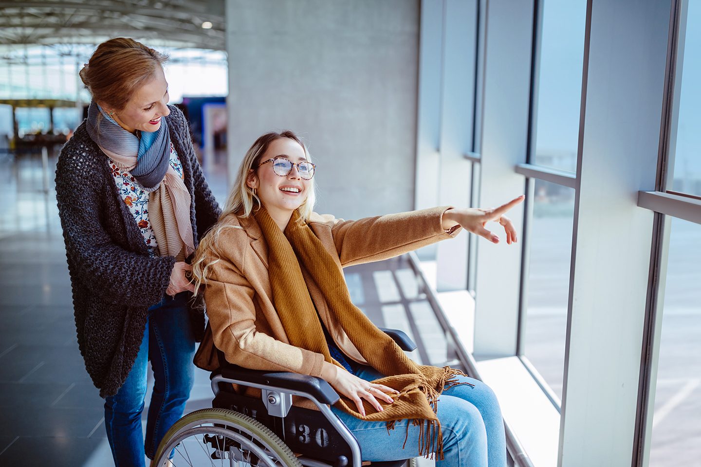 Young disabled woman on wheelchair and carer waiting at airport and looking at window view