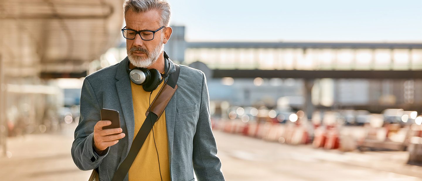 Mature man using mobile phone while walking on sidewalk. Confident male commuter holding smartphone in city. He is wearing blazer.