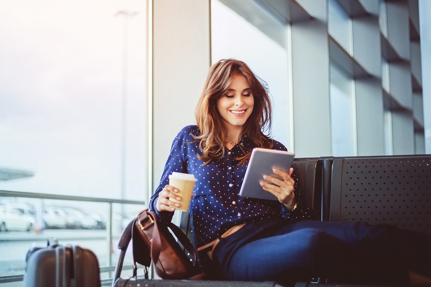 Shot of happy young woman with coffee waiting her flight at airport lounge and reading ebook on her digital tablet.