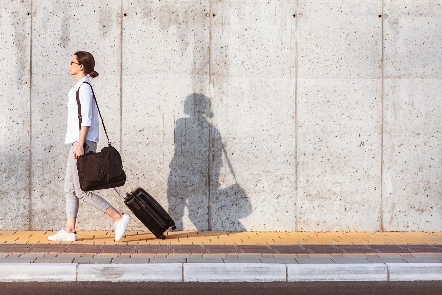 Attractive young woman walking on a sidewalk beside the concrete wall and pulling a small wheeled luggage. It seems that she is in a good mood. The shot is executed with available natural light, and the copy space has been left.